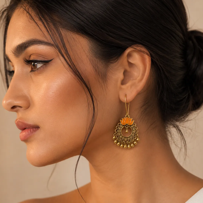A close-up side profile of a woman wearing a dangling gold-toned earring with an orange lotus flower design and small gold beads at the bottom.