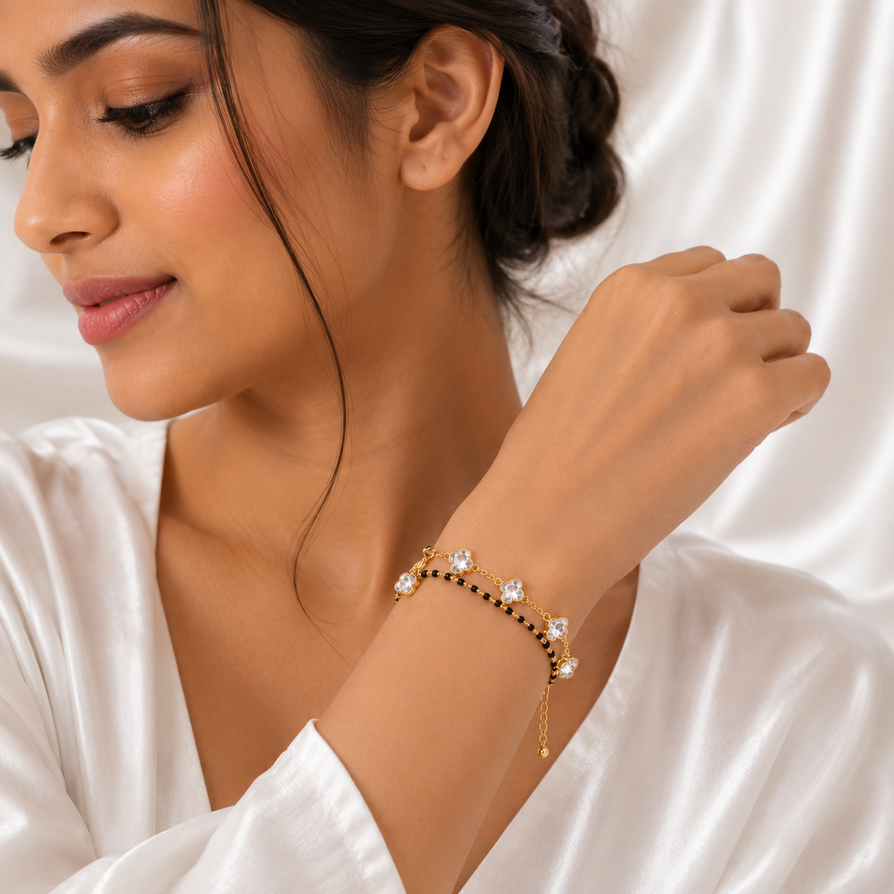 A woman wearing a delicate gold bracelet with clear crystal-like beads and small black accents, photographed against a soft white satin background.
