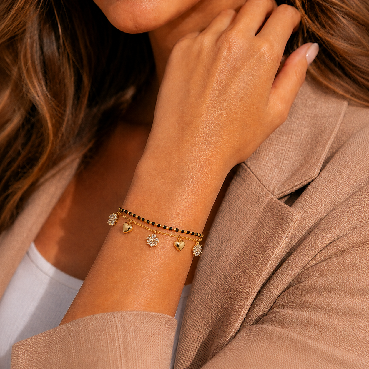 A close-up of a woman wearing a delicate gold bracelet with heart charms and small sunburst details, paired with a black beaded bracelet, on her wrist.