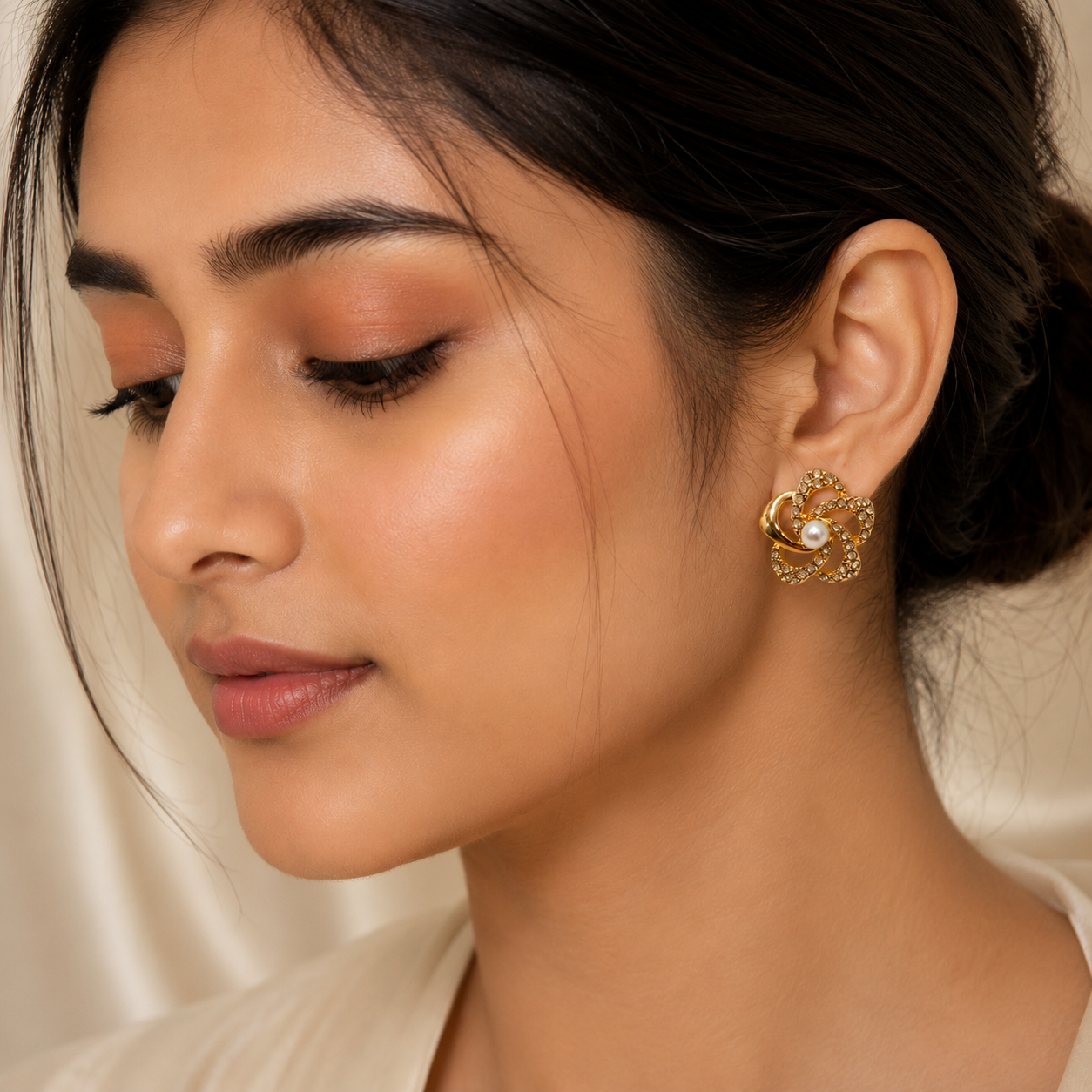A close-up of a woman wearing a gold-toned, flower-like earring with a central pearl, set against a soft cream satin background.