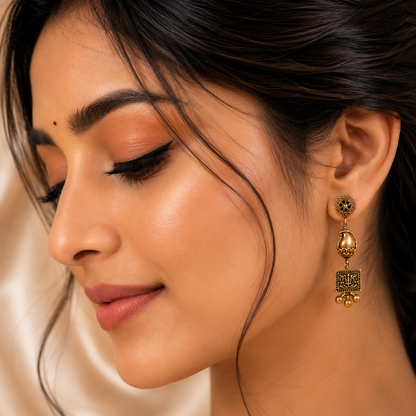 A close-up of a woman wearing a gold-toned traditional Indian jhumka earring with a square filigree charm and a small gold bell at the bottom.