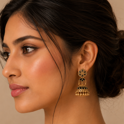A close-up side profile of a woman wearing a gold-toned traditional Indian jhumka earring with black enamel detailing, set against a warm beige background.