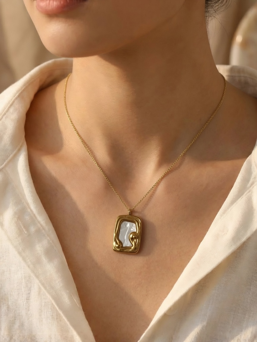 A close-up of a woman wearing a gold tonned chain necklace with an abstract rectangular pendant featuring a white inlay, styled with a white linen shirt.