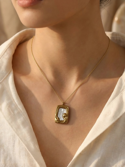A close-up of a woman wearing a gold tonned chain necklace with an abstract rectangular pendant featuring a white inlay, styled with a white linen shirt.