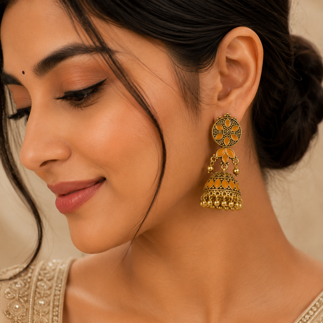 A close-up of a woman wearing a large, ornate gold-toned jhumka earring with intricate black and yellow stone detailing, set against a soft beige background.