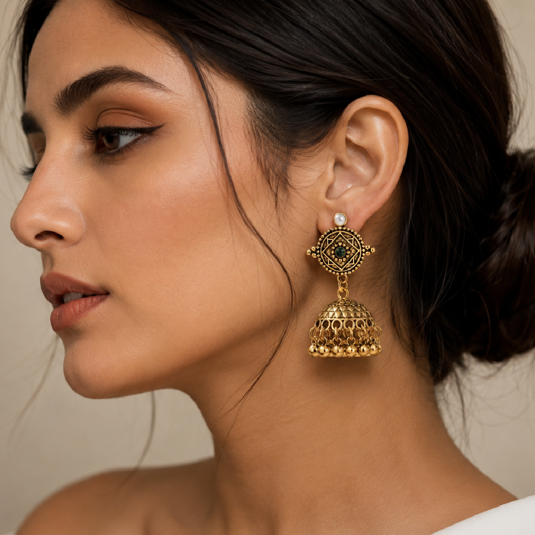 A close-up side profile of a woman wearing a large ornate gold jhumka earring with a small white stone accent on the post, against a soft beige background.