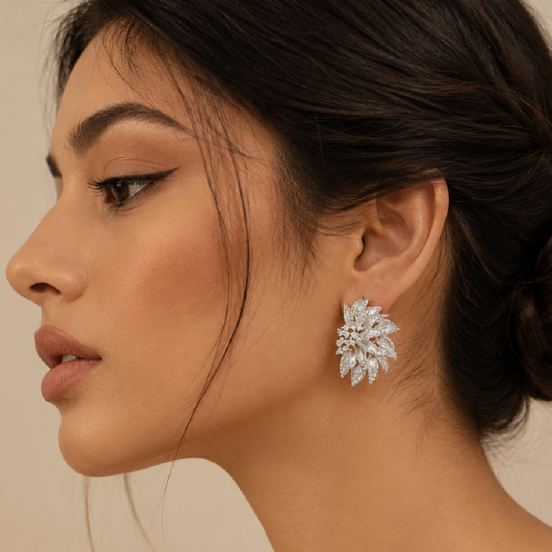 A close-up side profile of a woman wearing a silver-toned, leafy floral stud earring with clear stones against a warm beige background.