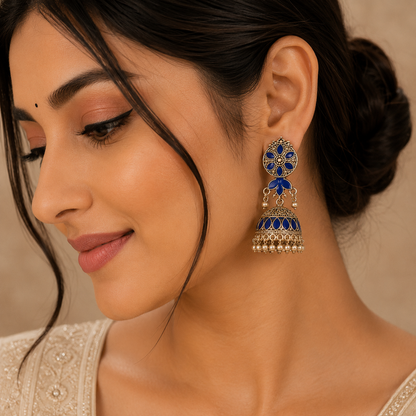A close-up of a woman wearing a single ornate gold-toned jhumka earring with deep blue enamel detailing and small clear stones, shown against a soft beige background.