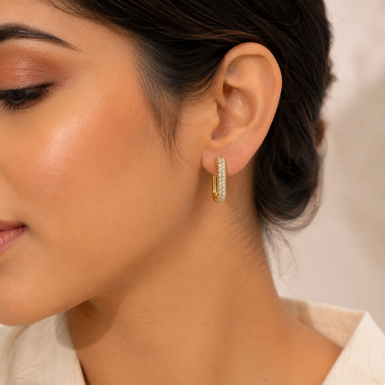 A close-up of a woman's ear wearing a small gold huggie hoop earring adorned with clear pavé stones. The background is a soft, warm beige tone.