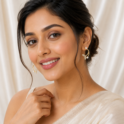 A close-up portrait of a woman wearing gold tonned crescent-shaped earrings with small white pearl-like accents, set against a soft cream-colored draped fabric background.