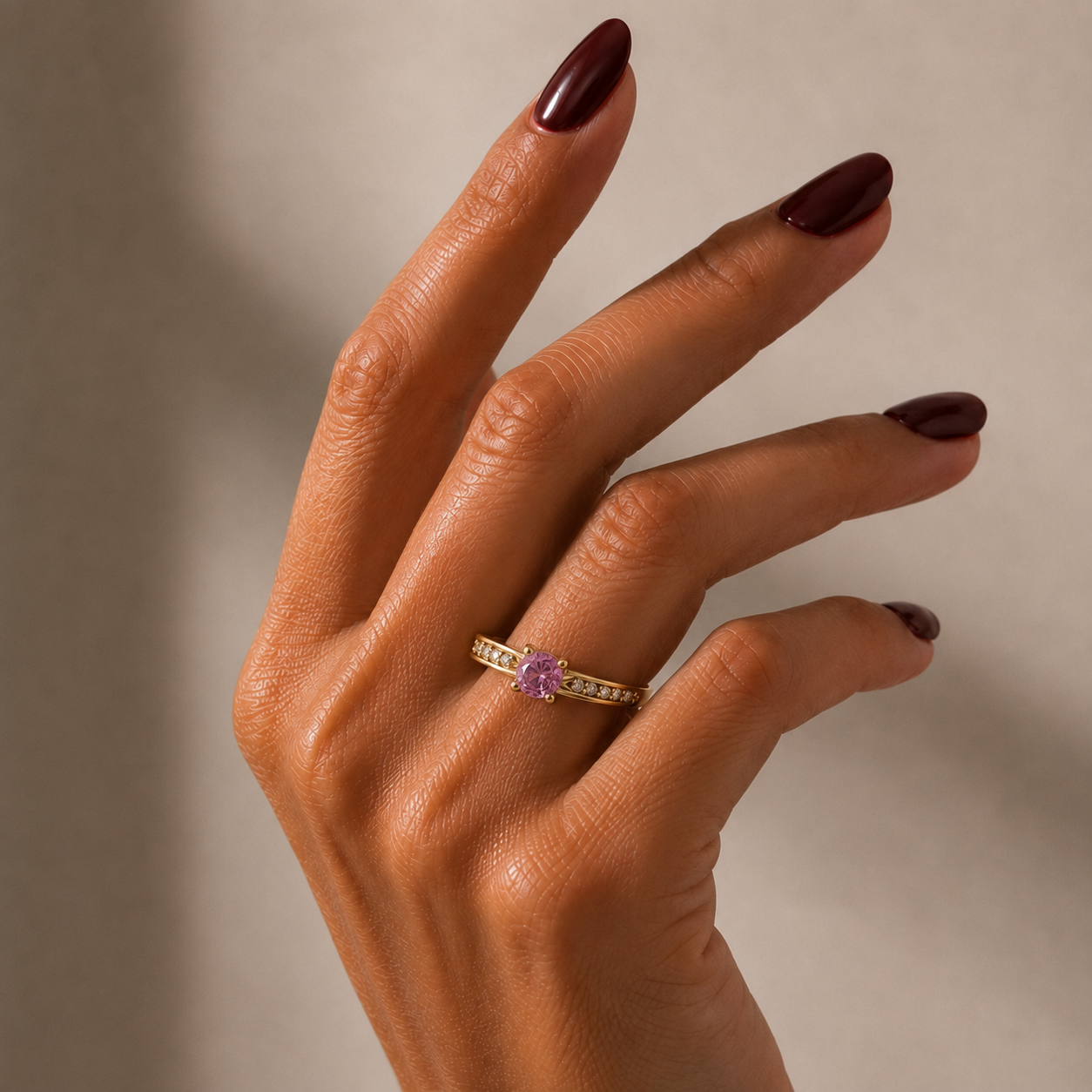 A close-up of a hand with dark burgundy manicured nails wearing a yellow gold-tonned ring with a central purple stone and small clear accent stones.